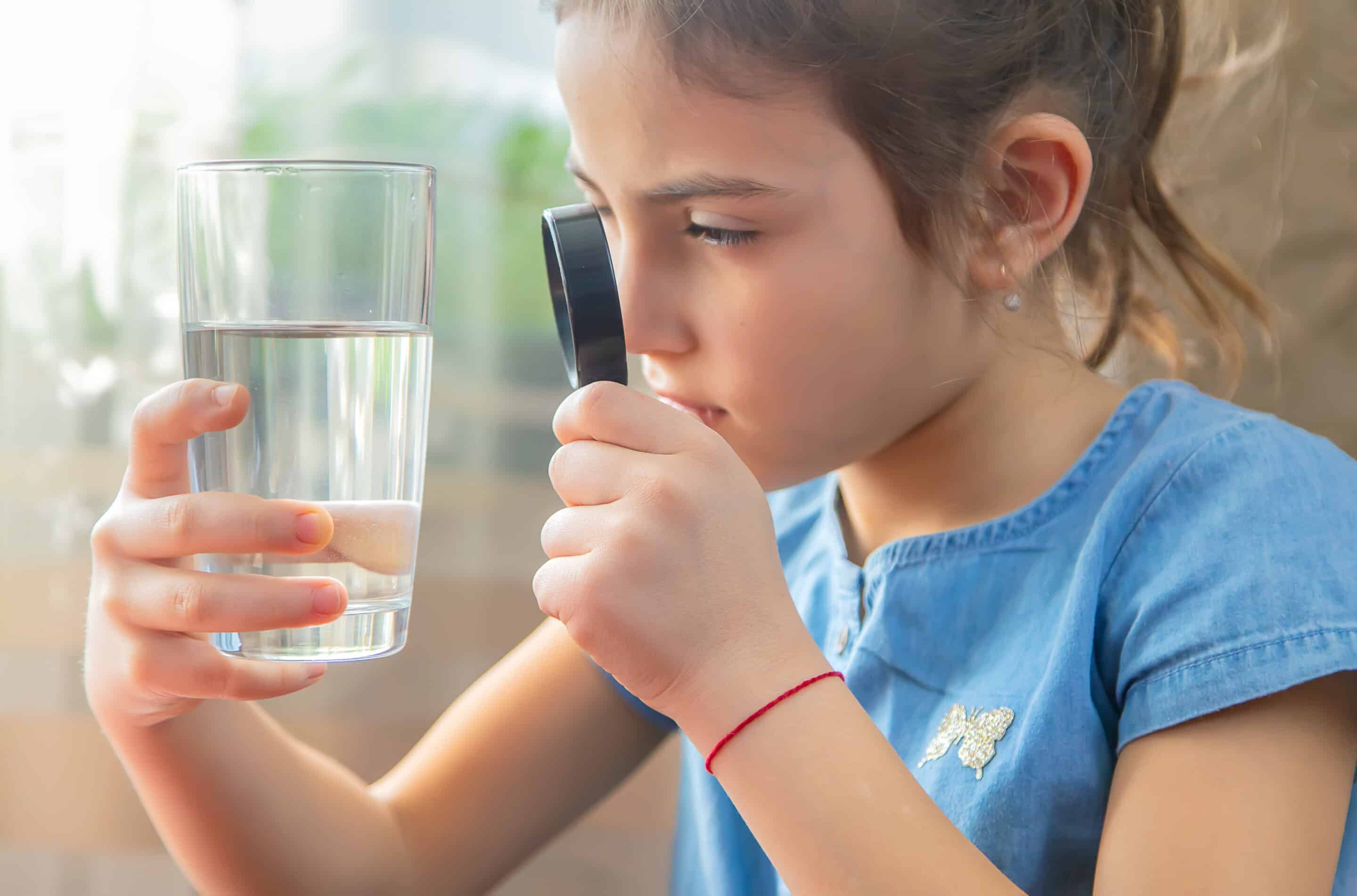 Girl inspecting glass of drinking water through microscope; radon in water poses health risks.